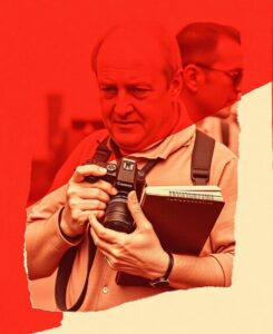 Older man holds a Canon camera and a spiral notebook, focused on taking a photo, with a red-toned background.